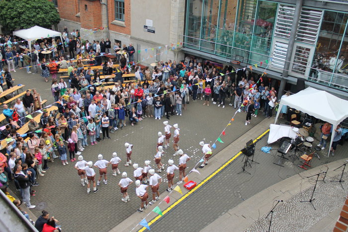 Ein Bild von oben auf den Hof der Schule, während eines Auftritts von Schülerinnen und Schülern mit weißen Mützen, T-Shirt und Lederhosen. Viele andere Personen schauen zu