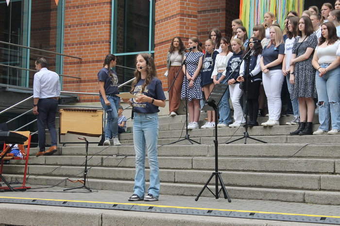 Eine Schülerin spricht in ein Mikrofon und hält Zettel in der Hand. Hinter ihre stehen Schülerinnen und Schüler auf Treppenstufen