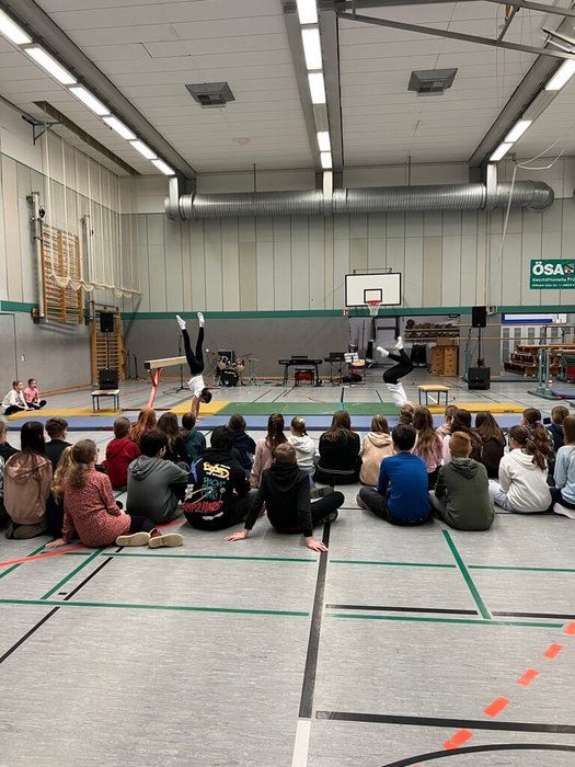 Zwei Schülerinnen turnen auf Matten und sind von anderen Schülerinnen und Schülern umgeben, die zuschauen