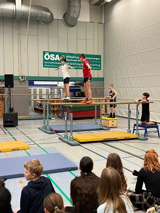 Zwei Schüler turnen an zwei Barren und zwei andere Schüler sitzen an den Enden eines jeden Barrens 