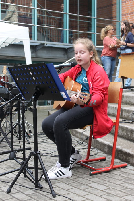 Eine Schülerin sitzt auf einem Stuhl, singt und spielt Gitarre. Sie trägt eine rote Jacke und eine schwarze Hose