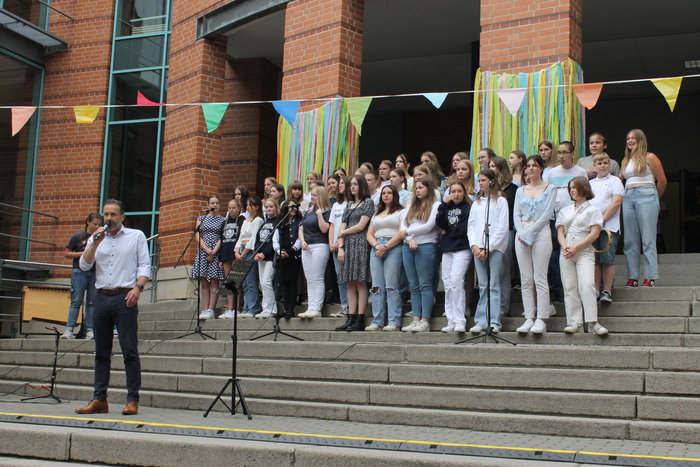 Schuldirektor Pasderski spricht ins Mikrofon. Auf den Treppnstufen dahinter stehen Schülerinnen und Schüler